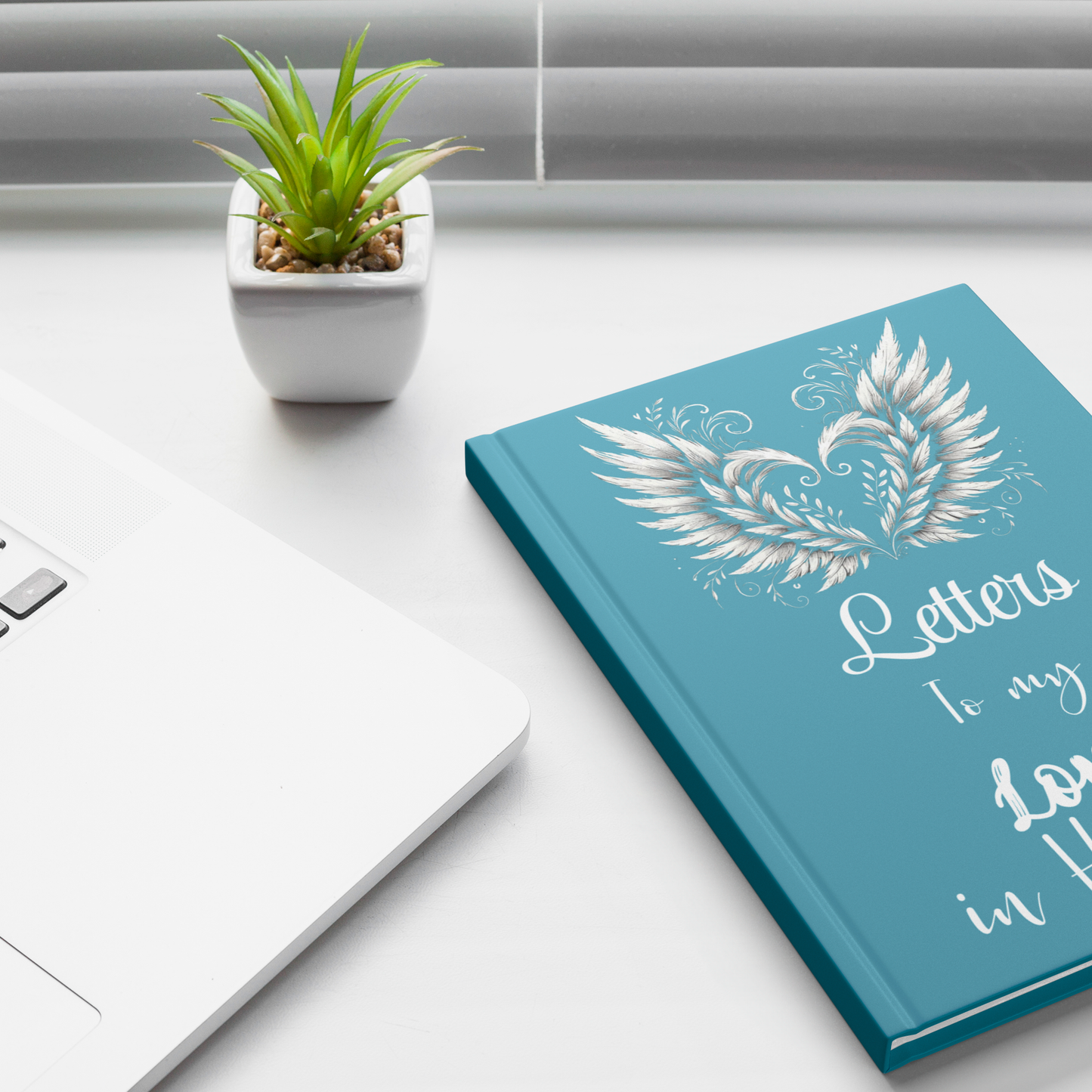 Teal hardcover notebook with silver angel wings and embossed "Letters to my Love in Heaven" text on white desk.