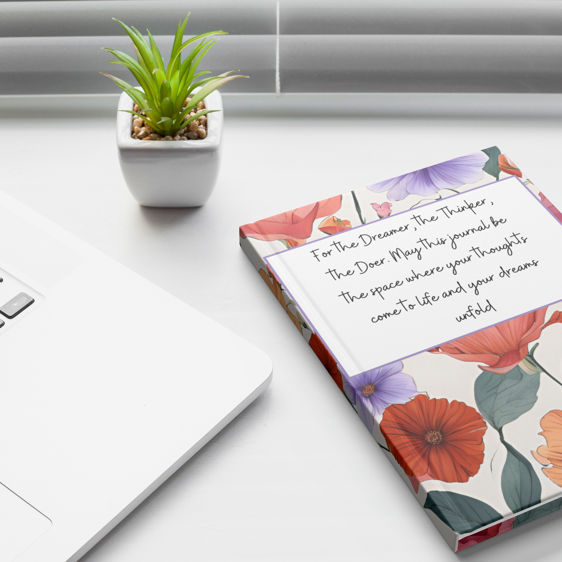 Floral notebook with handwritten motivational quote on white desk beside silver laptop and small green plant in white pot.