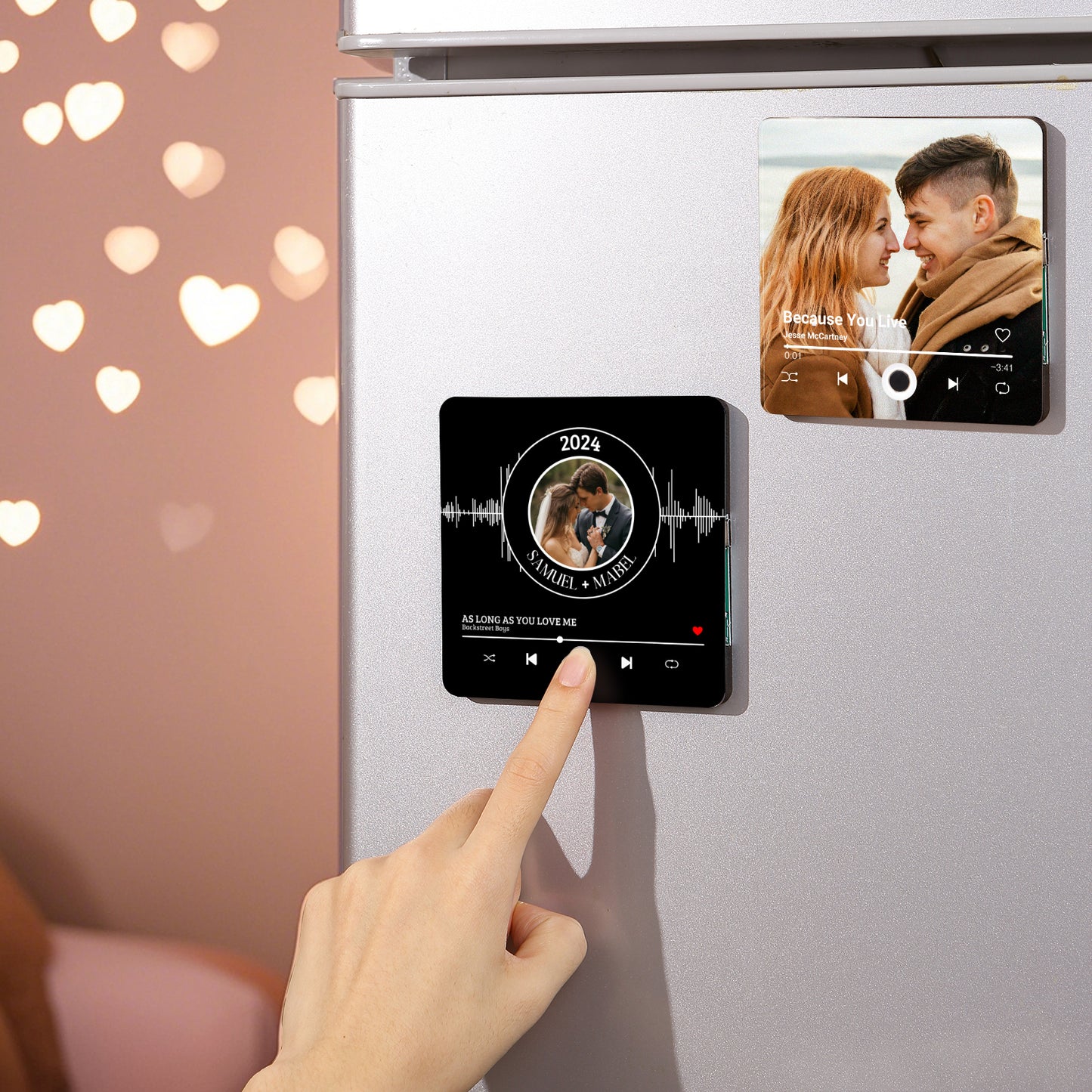 Close-up of a person's hand touching a black square magnet on a refrigerator, featuring a couple's photo and music player controls, with another magnet above it displaying a couple's photo and "Because You Live" by Jesse McCartney.