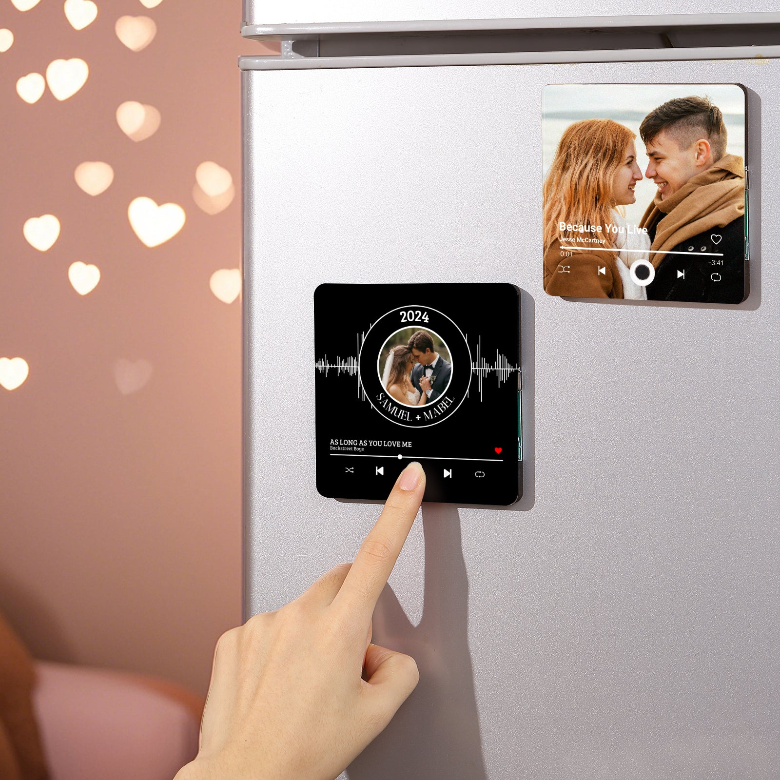 Close-up of a person's hand touching a black square magnet on a refrigerator, featuring a couple's photo and music player controls, with another magnet above it displaying a couple's photo and "Because You Live" by Jesse McCartney.